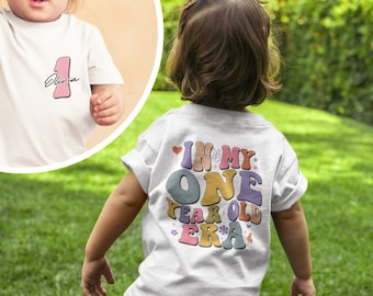 May include: A white toddler t-shirt with a colorful graphic that says "In My One Year Old Era". The shirt is being worn by a toddler in a grassy field.