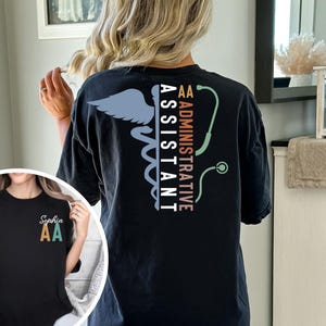 May include: Black t-shirt with a medical-themed design. The back features a Caduceus symbol, the words "ADMINISTRATIVE ASSISTANT", and a stethoscope graphic. The front of the shirt has the initials "AA" and the name "Sophia".