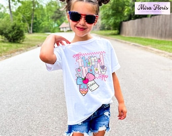 Op de afbeelding: Een wit t-shirt met een kleurrijke afbeelding met de tekst "Third Grade" met een potlood, strik en bloemen. Het shirt wordt gedragen door een jong persoon met een zonnebril en een spijkerbroek.