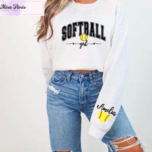 May include: White sweatshirt with the text "SOFTBALL girl" in black and yellow. The word "SOFTBALL" is arched across the top, with a yellow softball in the center. The sleeve has the name "Amelia" and a yellow softball.