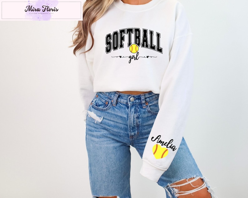 May include: White sweatshirt with the text "SOFTBALL girl" in black and yellow. The word "SOFTBALL" is arched across the top, with a yellow softball in the center. The sleeve has the name "Amelia" and a yellow softball.