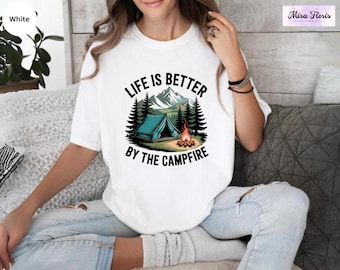 May include: White t-shirt with a camping graphic. The graphic features a teal tent, a campfire, trees, and mountains, with the text "LIFE IS BETTER BY THE CAMPFIRE" in black and green.