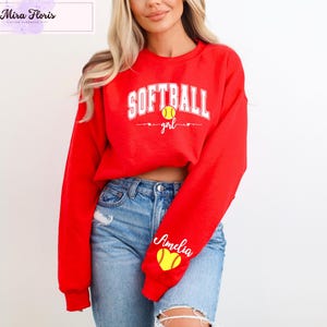 May include: A red sweatshirt with the words "SOFTBALL girl" in white, a yellow softball graphic, and the name "Amelia" on the sleeve. The sweatshirt is paired with blue jeans.