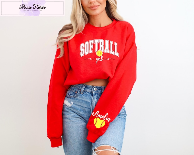 May include: A red sweatshirt with the words "SOFTBALL girl" in white, a yellow softball graphic, and the name "Amelia" on the sleeve. The sweatshirt is paired with blue jeans.