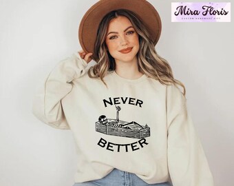 May include: Beige sweatshirt with the text "NEVER BETTER" in black, featuring a skeleton in a coffin. The sweatshirt is a long-sleeved crew neck. The Mira Floris logo is in the upper right corner.