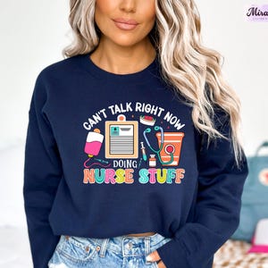 May include: A navy blue sweatshirt with the text "Can't talk right now doing nurse stuff" in rainbow colors. The text is surrounded by a stethoscope, a blood bag, a syringe, a clipboard, and a cup.