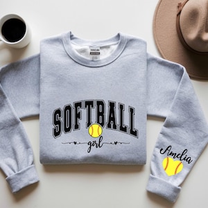 May include: A light gray sweatshirt with the word "SOFTBALL" in black block letters and a yellow softball graphic. The word "girl" is written below the softball. The right sleeve has the name "Amelia" and a softball graphic.