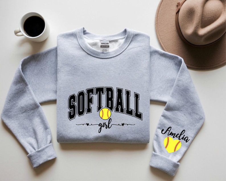 May include: A light gray sweatshirt with the word "SOFTBALL" in black block letters and a yellow softball graphic. The word "girl" is written below the softball. The right sleeve has the name "Amelia" and a softball graphic.