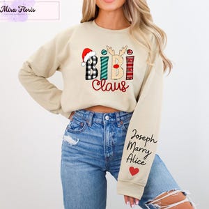 May include: Beige sweatshirt with a festive "Bibi Claus" design featuring a Santa hat, reindeer antlers, and plaid patterns. The right sleeve has names and a red heart. A Mira Floris logo is in the upper left corner.
