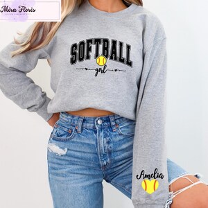 May include: A light gray sweatshirt with the words "SOFTBALL girl" in black and a yellow softball graphic. The name "Amelia" is printed on the sleeve with a yellow softball graphic. The sweatshirt is made of a soft material.