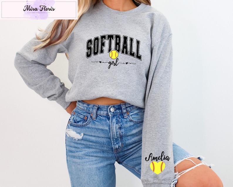 May include: A light gray sweatshirt with the words "SOFTBALL girl" in black and a yellow softball graphic. The name "Amelia" is printed on the sleeve with a yellow softball graphic. The sweatshirt is made of a soft material.