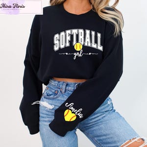May include: Black long-sleeve sweatshirt with the text "SOFTBALL girl" in white. A yellow softball graphic is centered above the word "girl". The sleeve features a yellow heart with a softball design and the name "Amelia" in script.