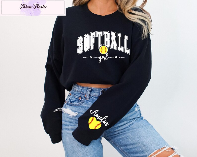 May include: Black long-sleeve sweatshirt with the text "SOFTBALL girl" in white. A yellow softball graphic is centered above the word "girl". The sleeve features a yellow heart with a softball design and the name "Amelia" in script.