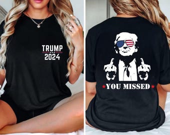 May include: Black t-shirt with white text that says "TRUMP 2024" on the front and a white image of a man wearing sunglasses with an American flag design and giving the middle finger on the back. The text on the back says "YOU MISSED".