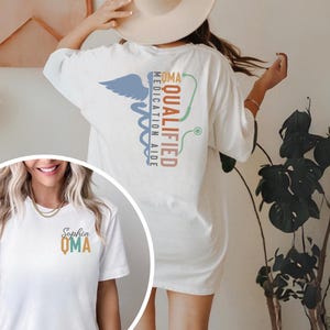 May include: White t-shirt with a medical caduceus design in blue, green, and brown. The text on the back reads "QMA Medication Aide Qualified." A smaller version of the design is on the front, with the name "Sophia" above "QMA."