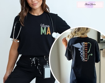 May include: Black t-shirt with a pocket on the front that says "Olivia MA" in green, orange, and white text. The back of the shirt has a medical symbol with the text "MA CERTIFIED MEDICAL ASSISTANT" in green, orange, and white text.