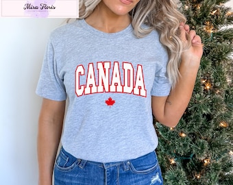 May include: A light gray t-shirt with the word "CANADA" in bold, red, block letters. Below the word is a red maple leaf. The shirt is made of a soft material.