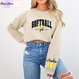 May include: A beige sweatshirt with the words "SOFTBALL girl" in black lettering and a yellow softball graphic. The sleeve features the name "Amelia" and a softball graphic. The sweatshirt is paired with blue jeans.