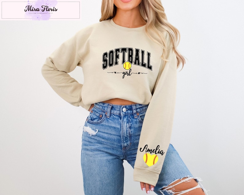 May include: A beige sweatshirt with the words "SOFTBALL girl" in black lettering and a yellow softball graphic. The sleeve features the name "Amelia" and a softball graphic. The sweatshirt is paired with blue jeans.