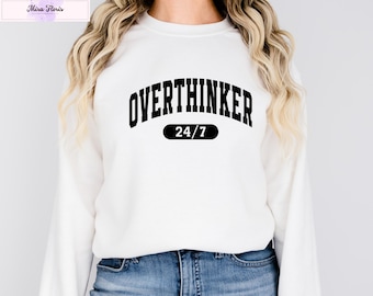 May include: White sweatshirt with the text "OVERTHINKER 24/7" in black, arched lettering. The text is centered on the chest. The sweatshirt has long sleeves and is worn with blue jeans.