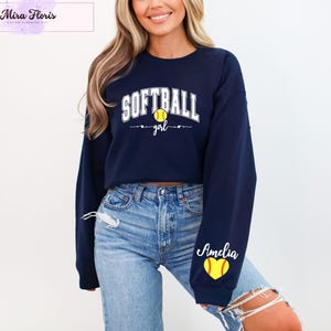May include: Navy blue sweatshirt with the word "SOFTBALL" in white block letters and a yellow softball graphic. The word "girl" is below the softball. The name "Amelia" is on the sleeve with a yellow softball graphic.