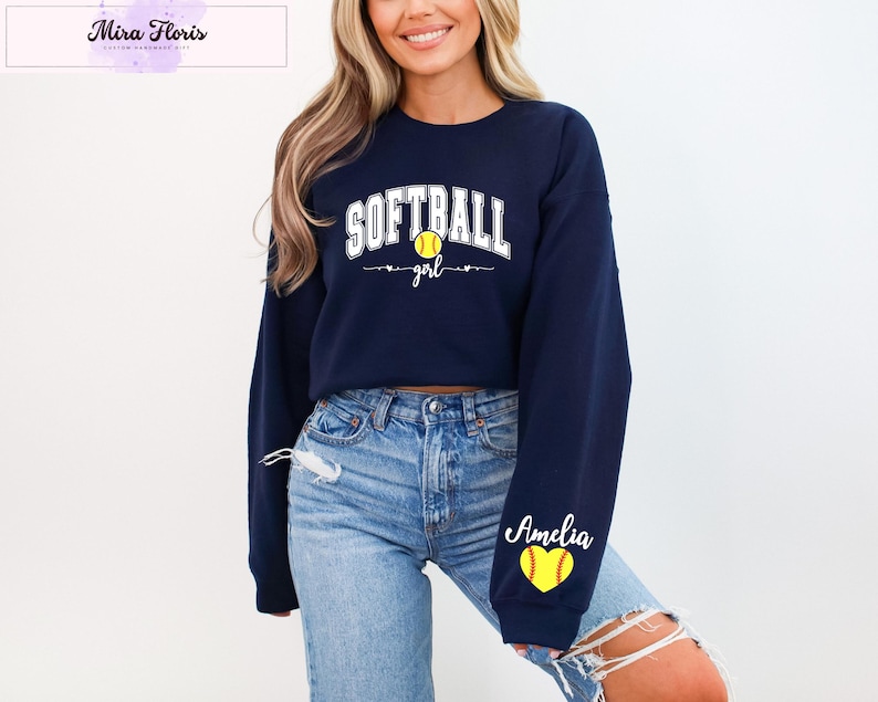 May include: Navy blue sweatshirt with the word "SOFTBALL" in white block letters and a yellow softball graphic. The word "girl" is below the softball. The name "Amelia" is on the sleeve with a yellow softball graphic.