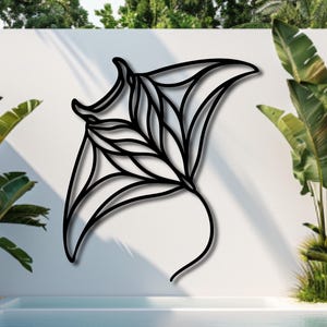 May include: A black metal wall art piece in the shape of a manta ray, mounted on a white wall. The artwork features intricate cut-out details and is displayed near a swimming pool and lush greenery.