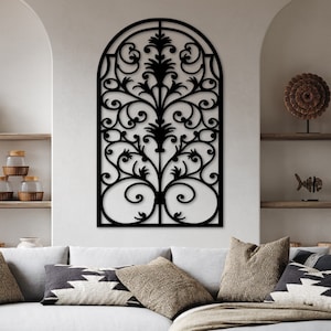 Op de afbeelding: Een zwarte metalen wanddecoratie met een decoratief boogontwerp en ingewikkelde krulwerk. De decoratie hangt aan een witte muur boven een lichtgrijze hoekbank met kussens.