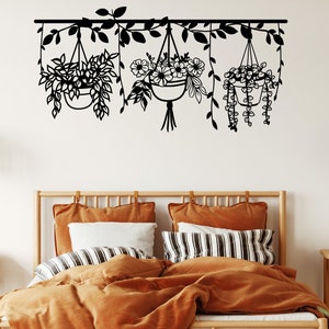May include: Black metal wall decor featuring three hanging plant pots with floral and leafy designs. The decor is mounted above a wooden bed frame with orange bedding and pillows.