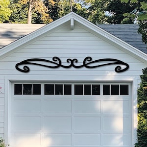 May include: A black metal scrollwork design mounted above a white garage door. The decorative piece features a symmetrical, flowing pattern with curved lines and swirls, adding a touch of elegance to the exterior.
