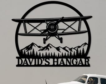 May include: Black metal wall art featuring a biplane design within a circular frame, mountains, and trees. The text "DAVID'S HANGAR" is displayed below. A white airplane is parked in front of the wall art.