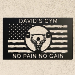 Peut inclure: Décoration murale en métal noir représentant un haltérophile soulevant une barre dans un cercle. Le design intègre un motif de drapeau américain avec le texte "DAVID'S GYM" en haut et "NO PAIN NO GAIN" en bas.