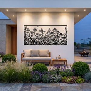 May include: A rectangular black metal wall art piece featuring a detailed laser-cut silhouette of a hummingbird hovering among various wildflowers, including daisies and dandelion seed heads, mounted on a plain white exterior wall.