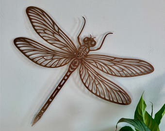 May include: Copper-colored metal dragonfly wall art. The dragonfly has detailed wing patterns and a slender body. The wall decor is mounted on a white wall, with green foliage in the lower right corner. The text "Color: Copper" is visible.