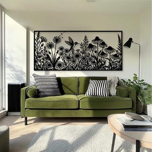May include: A rectangular black metal wall art piece featuring a detailed laser-cut silhouette of a hummingbird hovering among various wildflowers and dandelion seed heads, mounted horizontally on a plain white wall above a green sofa.