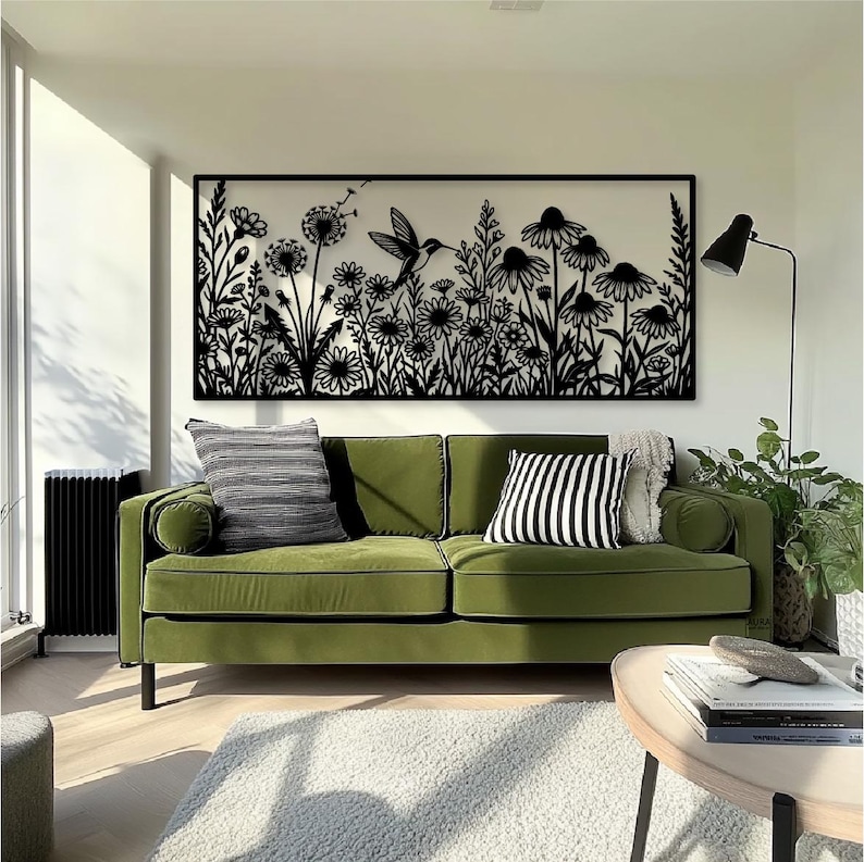 May include: A rectangular black metal wall art piece featuring a detailed laser-cut silhouette of a hummingbird hovering among various wildflowers and dandelion seed heads, mounted horizontally on a plain white wall above a green sofa.