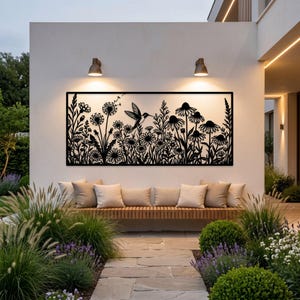 May include: A rectangular black metal wall art panel featuring a detailed laser-cut silhouette of a hummingbird hovering among various wildflowers, including daisies and dandelion seed heads, mounted on a white exterior wall above a bench.