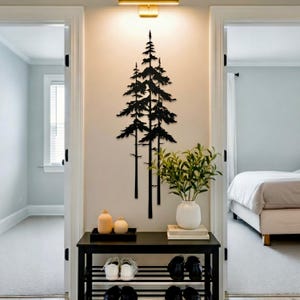 May include: Black metal tree wall art decor. The silhouette of three pine trees is mounted on a light-colored wall. A black shoe rack with shoes and decorative items sits below the artwork. A gold light fixture is above the artwork.