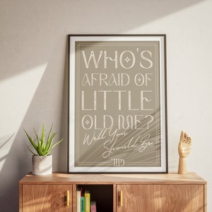 Peut inclure: Une impression encadrée avec un fond beige et un texte blanc qui dit "Who's Afraid of Little Old Rose? Well You Should Be." L'impression est accrochée à un mur au-dessus d'une étagère en bois avec une plante en pot et une sculpture de main en bois.