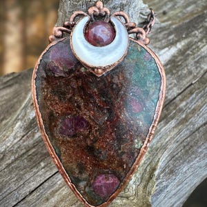 May include: A copper pendant with a heart-shaped green and brown stone with red flecks. The stone is set in a copper frame with a crescent moon design and a white shell inlay. The pendant is on a copper chain.