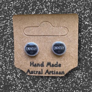 May include: A pair of black stud earrings with the word "EMPATHY" in white lettering on each earring. The earrings are displayed on a brown card with the text "Hand Made Astral Artisan".