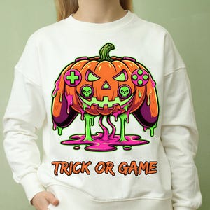 Op de afbeelding: Witte sweatshirt met een Halloween-thema. Het ontwerp bevat een oranje pompoen met een controllerontwerp, groene en paarse druipende accenten en de tekst "TRICK OR GAME" in oranje.