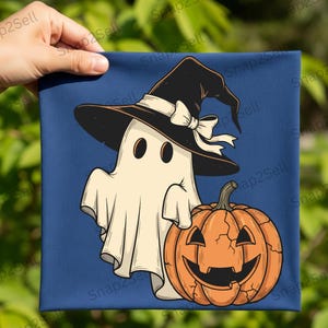 May include: A Halloween-themed canvas print featuring a cartoon ghost wearing a witch hat with a bow, and a carved orange pumpkin. The background is a solid dark blue. The print is held by a hand.