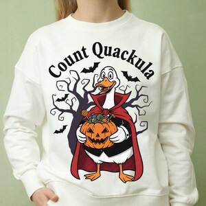 Op de afbeelding: Witte sweatshirt met een Halloween-thema. Het ontwerp toont een cartoon eend verkleed als een vampier, met een pompoen vol snoep. De tekst "Count Quackula" staat boven de eend, met vleermuizen en een enge boom op de achtergrond.