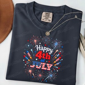 May include: A dark gray crew neck t-shirt featuring a graphic print of colorful fireworks and rockets surrounding the text Happy 4th of JULY, with the word JULY styled in red, white, and blue American flag stripes. The shirt has a Comfort Colors label.