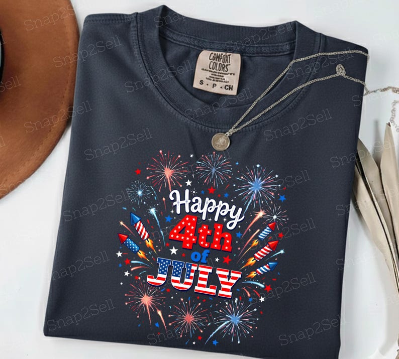 May include: A dark gray crew neck t-shirt featuring a graphic print of colorful fireworks and rockets surrounding the text Happy 4th of JULY, with the word JULY styled in red, white, and blue American flag stripes. The shirt has a Comfort Colors label.