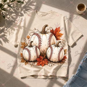 Könnte beinhalten: Ein cremefarbenes Kurzarm-T-Shirt mit einem Grafikdruck von drei weißen Kürbissen, die mit roten Nähten im Baseball-Stil versehen sind. Die Kürbisse sind von orangefarbenen und braunen Herbstblättern sowie dünnen Ranken umgeben.