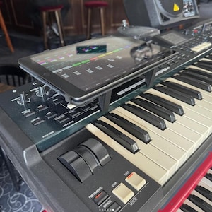 May include: A black keyboard with white keys and a tablet attached to the top. The tablet is displaying a music production app. The keyboard has a variety of knobs and buttons.
