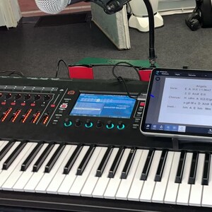 Könnte beinhalten: Eine schwarze Tastatur mit einem digitalen Display und einem Tablet, auf dem Notenblätter mit dem Titel "Angela" angezeigt werden. Die Notenblätter enthalten den Text "Verse 1, 2, Chorus, Intro, A, B, C, D, E, F, G, H, I, J, K, L, M, N, O, P, Q, R, S, T, U, V, W, X, Y, Z, 1, 2, 3, 4, 5, 6, 7, 8, 9, 0, und a, b, c, d, e, f, g, h, i, j, k, l, m, n, o, p, q, r, s, t, u, v, w, x, y, z".