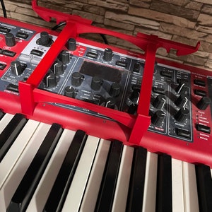 May include: A red synthesizer with a black and white keyboard and a red metal stand. The synthesizer has a variety of knobs and buttons, including a large display screen. The text "SYNTH" is visible on the synthesizer.